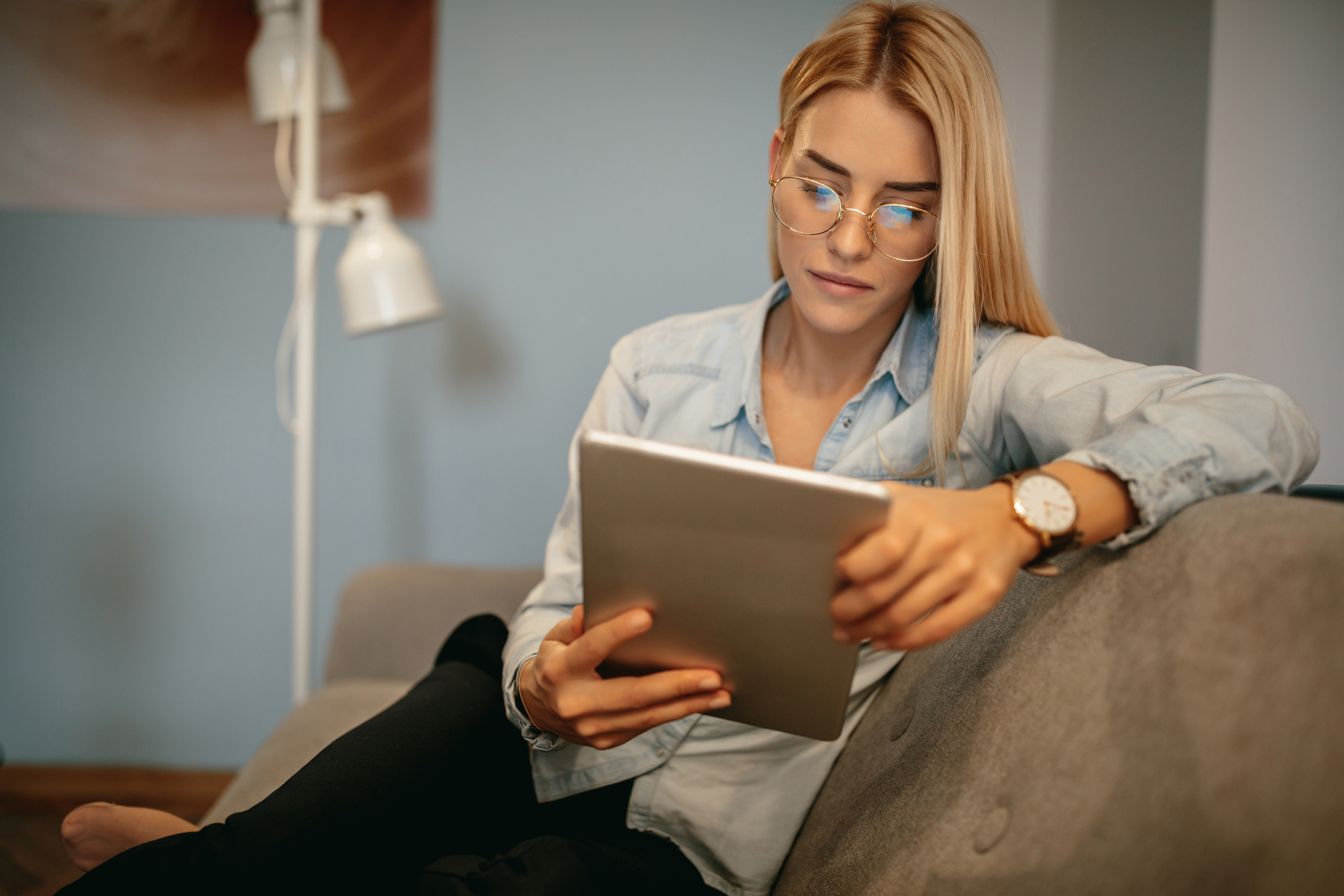 a white female sitting on the couch with her tablet reading something
